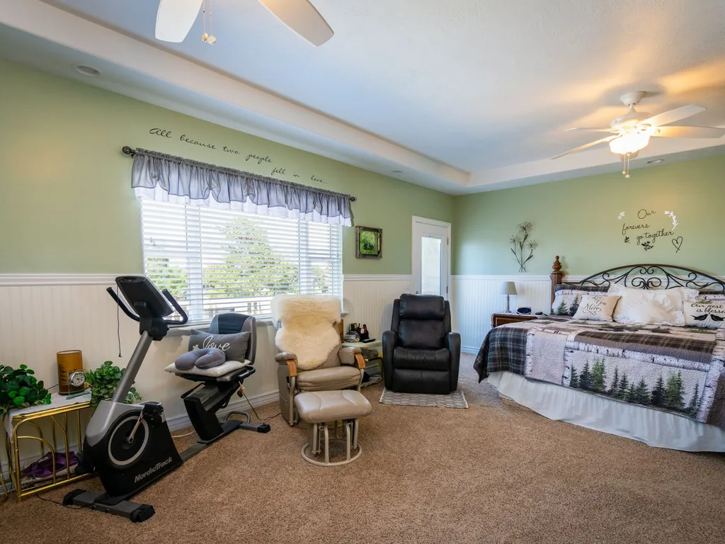 Real cluttered bedroom before AI room redesign — exercise bike, recliner, glider and ottoman, green walls, two ceiling fans and a tray ceiling